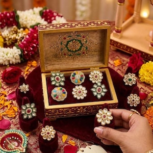 May include: A wooden jewellery box with a hinged lid featuring gold and green floral patterns, containing six rings with colourful stones and gold-tone metal settings. Additional rings are displayed on velvet stands around the box.