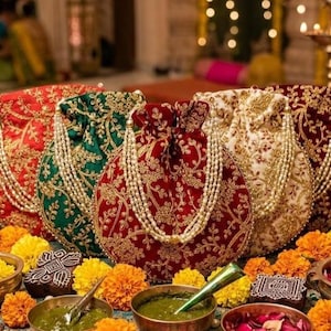 May include: Five round drawstring pouches in red, green, maroon, and white, each featuring intricate gold embroidery and a decorative pearl bead handle. The pouches are arranged in a row, surrounded by small bowls of marigold flowers and green paste.