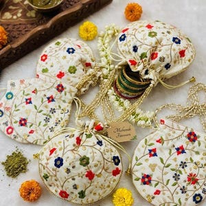 Puede incluir: Se muestran varias bolsas blancas bordadas con cordón y diseños florales en rojo, azul y verde. Las bolsas están rodeadas de flores de caléndula, henna y una bandeja de madera. Una etiqueta dice "Mehndi Favours".
