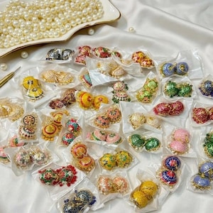 Puede incluir: Una colección de numerosos pendientes jhumka pequeños en forma de cúpula con diversos colores y patrones, empaquetados individualmente en plástico transparente, junto a una bandeja blanca con perlas blancas y un bolígrafo dorado.
