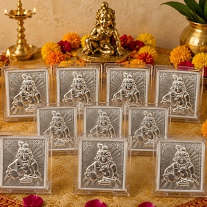 May include: A collection of nine square silver-toned plaques, each encased in a clear acrylic frame, featuring a raised relief of a seated deity figure with a sunburst background, arranged on a gold fabric surface with scattered flower petals.