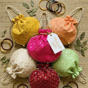 May include: A collection of colorful, embellished drawstring pouches in shades of yellow, pink, peach, cream, red, and green. Each pouch has a beaded handle and gold accents. The tag reads "Mehndi Gifts". Several bangles are also visible.