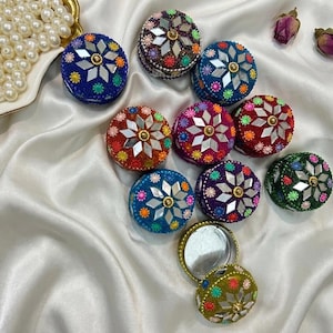 Puede incluir: Una colección de pequeños recipientes redondos con tapa que presentan espejos y bordados florales coloridos. Cada tapa muestra un patrón de espejo en forma de estrella rodeado de cuentas vibrantes. Un recipiente está abierto, revelando su interior.