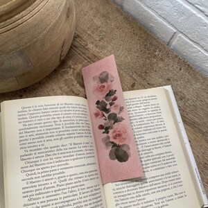 Può includere: Segnalibro rosa floreale con un design ad acquerello con rose e foglie. Il segnalibro rettangolare è posto su un libro aperto, aggiungendo un tocco di eleganza alla lettura.