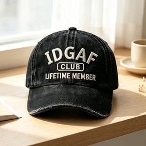 Op de afbeelding: Een zwarte baseballpet met een verwassen uiterlijk en wit geborduurde tekst IDGAF CLUB LIFETIME MEMBER. De pet heeft een gebogen klep, een gestructureerde kroon met een knoop bovenop en een zichtbare, doorleefde textuur in de stof.