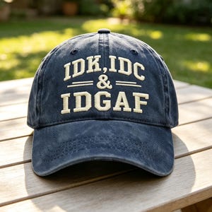 May include: A navy blue baseball cap with a distressed, washed-out finish. The front features embroidered cream-colored text that reads IDK, IDC & IDGAF in a bold, block-style font. The cap has a curved brim and a structured six-panel crown design.