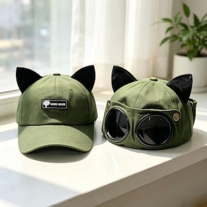 Könnte beinhalten: Zwei olivgrüne Baseballkappen mit schwarzen Katzenohren oben. Die linke Kappe hat einen rechteckigen schwarzen Aufnäher mit dem Text "HAND MADE" und einem Baumsymbol. Die rechte Kappe verfügt über integrierte dunkle, runde Schutzbrillen.