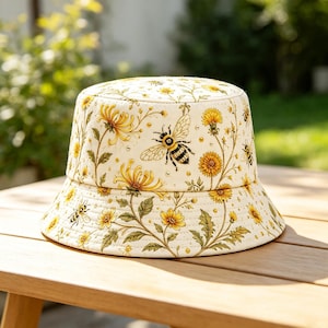 Może przedstawiać: Kremowy kapelusz typu bucket hat z powtarzającym się botanicznym nadrukiem przedstawiającym żółte kwiaty mniszka lekarskiego, zielone liście oraz czarno-żółte pszczoły. Wzór pokrywa całą główkę oraz opadające rondo kapelusza.