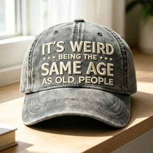 Può includere: Un cappellino da baseball grigio antracite con una finitura slavata e vissuta. La parte anteriore presenta un testo ricamato color crema che recita: IT'S WEIRD BEING THE SAME AGE AS OLD PEOPLE, con tre piccole stelle su ogni lato.