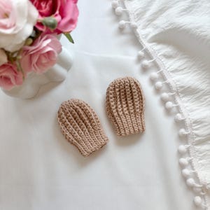 May include: A pair of light brown crochet baby mittens featuring a vertical ribbed texture. The mittens are crafted from yarn with a consistent stitch pattern, showing a rounded top and a slightly tapered shape at the base for a secure fit.