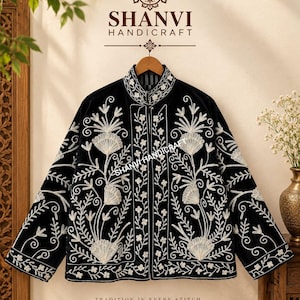 Puede incluir: Una chaqueta negra con bordados florales blancos, cuello alto y mangas largas. La prenda muestra el texto SHANVI HANDICRAFT en la parte superior e inferior, con el nombre de la marca integrado sutilmente en el centro del bordado.