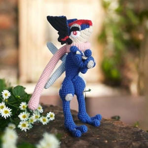 Puede incluir: Muñeca de ganchillo hecha a mano con forma de criatura azul y blanca con un ala negra, bufanda rosa y cabeza roja y blanca. La muñeca está sobre una superficie marrón con flores blancas en primer plano. La muñeca tiene brazos y piernas largos.