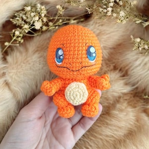 Puede incluir: Muñeca de ganchillo naranja hecha a mano con ojos azules y vientre color crema. La muñeca tiene brazos y piernas pequeños y una cara sonriente. La muñeca es un personaje Charmander de la popular serie Pokémon.