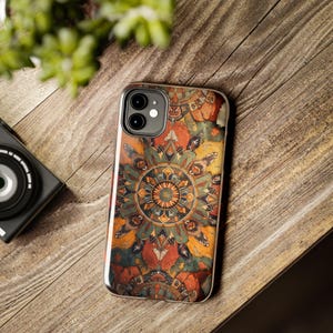 Pode incluir: Uma capa de smartphone com um padrão de mandala detalhado e simétrico em tons de laranja queimado, azul-petróleo profundo, amarelo mostarda e castanho escuro. O design cobre toda a parte de trás da capa, que tem uma borda transparente.
