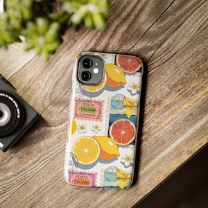 Pode incluir: Uma capa de telefone branca com um design vibrante de frutas e selos postais. A capa apresenta ilustrações de laranjas, limões e fatias de toranja, juntamente com detalhes florais e selos postais italianos. Sobre uma superfície de madeira.