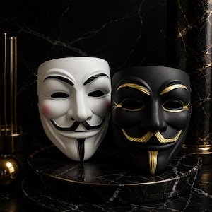 Puede incluir: Dos máscaras de Guy Fawkes, una blanca con detalles negros y mejillas rosadas, y otra negra mate con detalles dorados. Ambas máscaras presentan un bigote estilizado, perilla y cejas arqueadas, presentadas como un par decorativo.