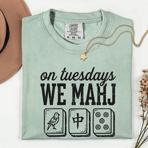 May include: A light sage green cotton t-shirt featuring the black text 'on tuesdays WE MAHJ' above three illustrations of mahjong tiles. The shirt has a Comfort Colors label at the neck and is paired with a gold star pendant necklace.