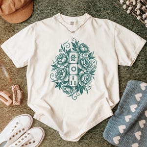 Puede incluir: Camiseta color crema con un diseño floral turquesa y caracteres chinos. El diseño incluye peonías y baldosas rectangulares. La camiseta es informal y tiene mangas cortas.