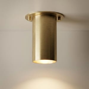 Op de afbeelding: Een cilindrische messing plafondlamp. De lamp heeft een ronde basis en een lange, buisvormige behuizing. Gemonteerd aan een wit plafond, straalt het een warm licht uit. Het ontwerp is minimalistisch en modern.