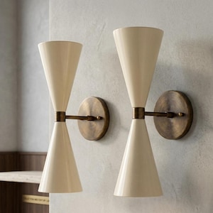 May include: A pair of wall sconces featuring dual off-white conical shades arranged in an hourglass shape. Each fixture is supported by a circular, antique brass-finished wall mount connected to the center of the cones by a horizontal metal arm.