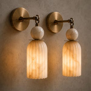 May include: Two wall sconces with a warm, inviting glow. Each sconce features a round, brass-colored wall plate, a curved arm, and a decorative, ribbed, off-white shade. The shades are topped with a white marble sphere and emit a soft light.