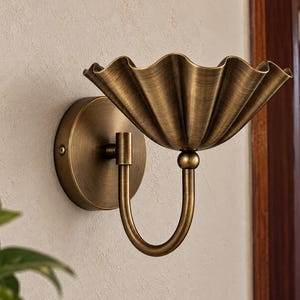 May include: A wall-mounted light fixture featuring a circular brass backplate and a curved arm supporting a wide, fluted, shell-shaped brass shade. The metal has a brushed, antique finish with visible texture and warm golden-brown tones.