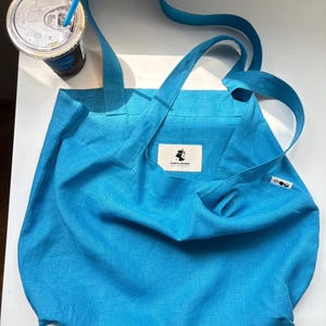 May include: A bright blue tote bag with long shoulder straps and a rectangular fabric label featuring a black graphic logo and the text LeeYu.Jukka. A small white side tag with a black circular icon is attached to the right edge of the bag.