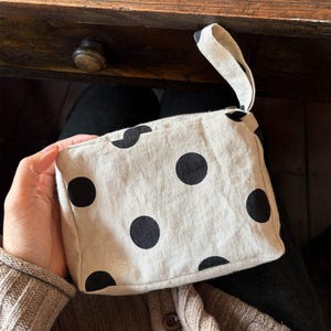 Puede incluir: Un estuche rectangular de tela beige con un patrón de grandes lunares negros. El estuche incluye una correa de muñeca de tela beige a juego unida a la esquina superior y un cierre de cremallera en el borde superior para guardar objetos.
