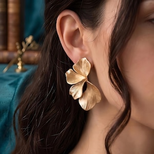 May include: Gold-toned, statement earrings in a floral design. The earrings feature three textured, petal-shaped elements, creating a layered, dimensional look. The earrings are worn on an ear, with the focus on the jewelry's design and color.