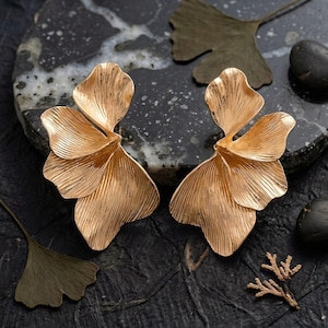 May include: A pair of gold-coloured, petal-shaped earrings. The earrings have a textured surface with detailed veining, resembling stylized flowers. The earrings are displayed on a dark, textured surface with decorative leaves and stones.
