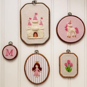 Puede incluir: Cinco tapices de lana fieltrada en bastidores de madera. Los diseños incluyen un castillo blanco con banderas rosas, un unicornio blanco con crin rosa, una letra M rosa, una figura con pelo castaño y vestido rosa, y tres tulipanes rosas con hojas verdes.