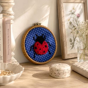 Pu&ograve; includere: Un telaio da ricamo circolare in legno contenente uno sfondo in tessuto blu a pois bianchi e una coccinella in lana infeltrita con un corpo rosso brillante a pois neri e una testa nera, centrata all'interno della cornice per l'esposizione.
