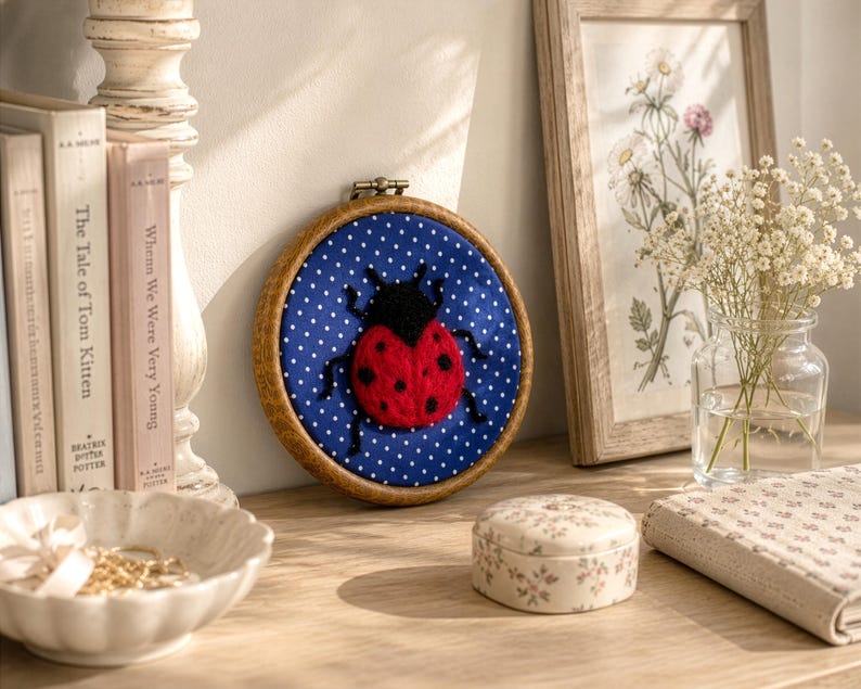 Pu&ograve; includere: Un telaio da ricamo circolare in legno contenente uno sfondo in tessuto blu a pois bianchi e una coccinella in lana infeltrita con un corpo rosso brillante a pois neri e una testa nera, centrata all'interno della cornice per l'esposizione.