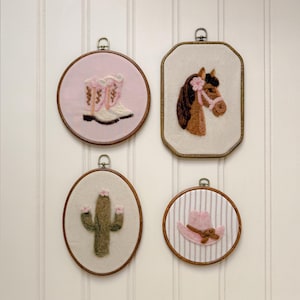 May include: Four wall-mounted embroidery hoops featuring needle-felted designs: a pair of white and pink cowboy boots on light pink fabric, a brown horse head on cream fabric, a green cactus on cream fabric, and a pink cowboy hat on striped fabric.