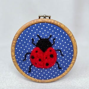 Pu&ograve; includere: Un telaio da ricamo circolare in legno contenente uno sfondo in tessuto blu con un motivo a pois bianchi. Al centro del tessuto si trova una coccinella in lana infeltrita con testa nera, corpo rosso brillante con punti neri e sei zampe.