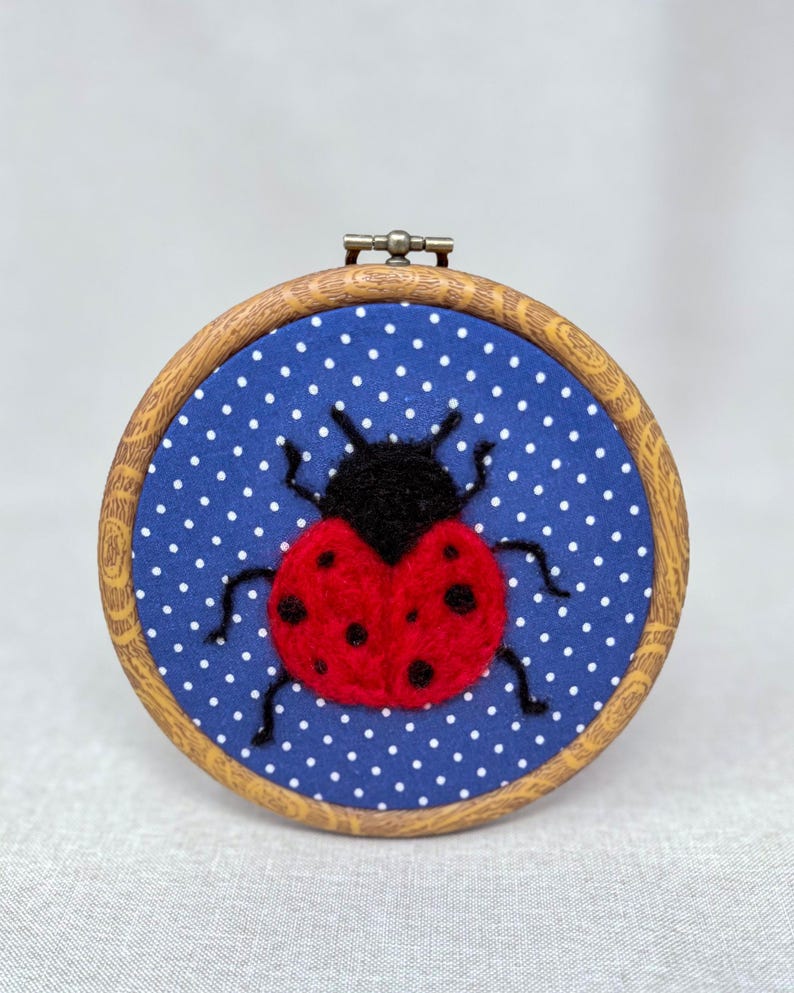 Pu&ograve; includere: Un telaio da ricamo circolare in legno contenente uno sfondo in tessuto blu con un motivo a pois bianchi. Al centro del tessuto si trova una coccinella in lana infeltrita con testa nera, corpo rosso brillante con punti neri e sei zampe.