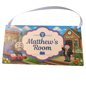 May include: A rectangular hanging sign with a white ribbon featuring a blue Thomas the Tank Engine, Sir Topham Hatt, and a train station. The centre has a scalloped white label with the text 'Matthew's Room' in blue, a yellow 'T' logo, and a small blue train icon.