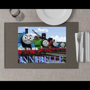 May include: A rectangular placemat featuring a printed scene of four colourful cartoon trains on railway tracks under a blue sky. The name ANNABELLE is printed in large, light blue and purple block letters across the bottom of the train graphic.