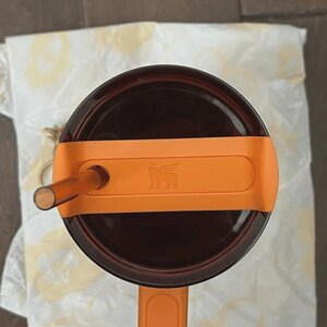 May include: An overhead view of a brown insulated tumbler with an orange handle and lid. The lid has a straw and an orange band with a logo. The tumbler sits on a white patterned cloth.