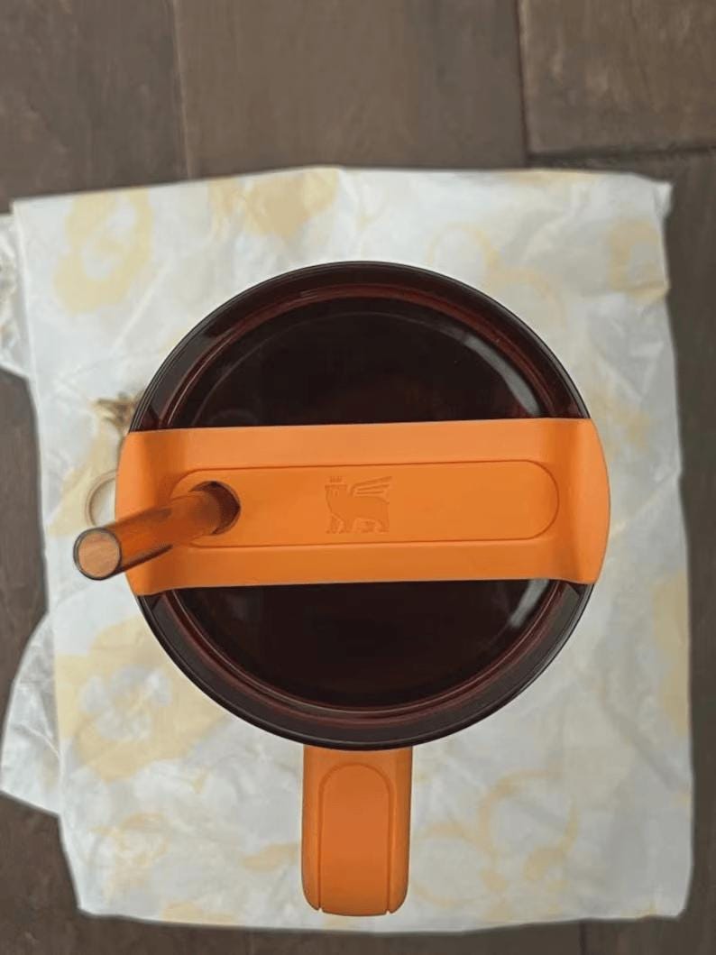 May include: An overhead view of a brown insulated tumbler with an orange handle and lid. The lid has a straw and an orange band with a logo. The tumbler sits on a white patterned cloth.
