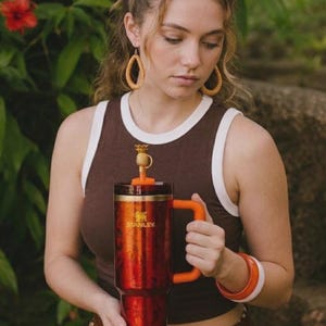 May include: A large, red and orange insulated tumbler with a handle. The tumbler has the brand name "STANLEY" printed on the side. It features a pineapple-shaped stopper and is held by a person wearing a brown tank top.