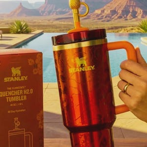 May include: A red and gold Stanley Quencher H2.0 tumbler with a pineapple-shaped topper. The tumbler has a handle and is 40 oz (1.18 L). The box next to it has the Stanley logo and the words "The Flonstate Quencher H2.0 Tumbler".