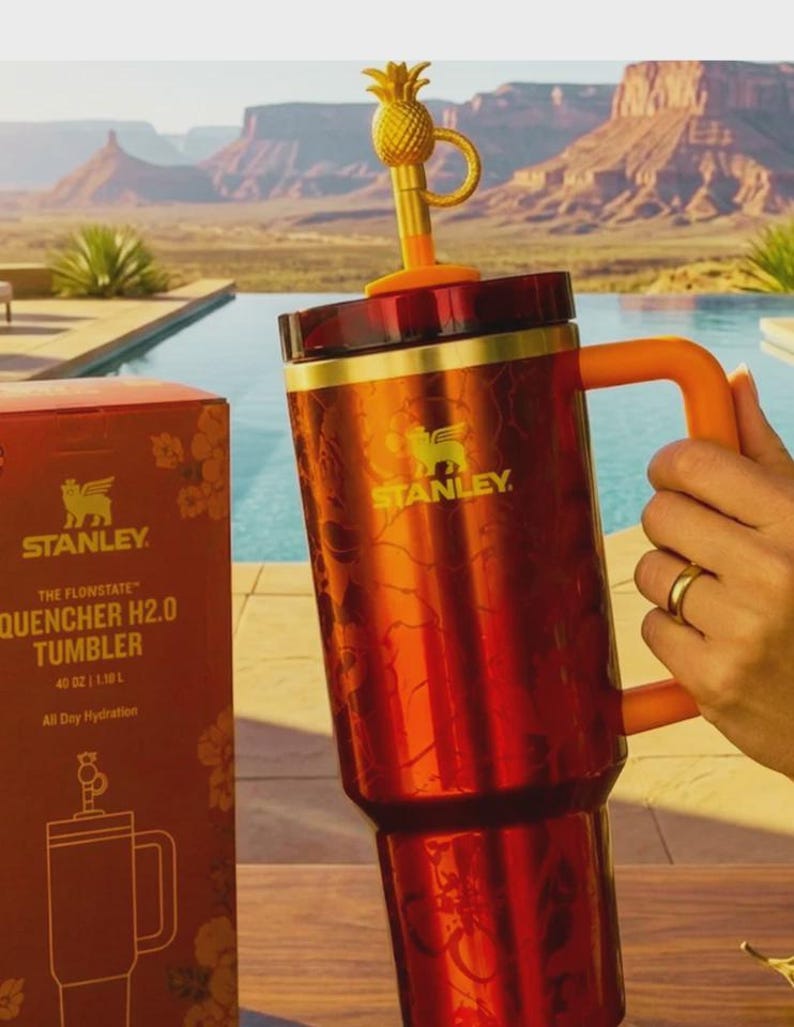 May include: A red and gold Stanley Quencher H2.0 tumbler with a pineapple-shaped topper. The tumbler has a handle and is 40 oz (1.18 L). The box next to it has the Stanley logo and the words "The Flonstate Quencher H2.0 Tumbler".