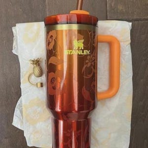 May include: A red and gold Stanley tumbler with a handle and straw. The tumbler has a floral design and the Stanley logo. An orange handle and straw complement the design. A gold pineapple charm is also present.