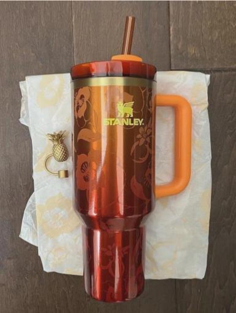 May include: A red and gold Stanley tumbler with a handle and straw. The tumbler has a floral design and the Stanley logo. An orange handle and straw complement the design. A gold pineapple charm is also present.