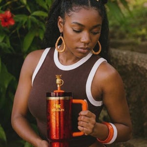 May include: A large, red and orange Stanley travel mug with a pineapple-shaped topper. The mug has a handle and the Stanley logo. The person in the image is wearing a brown tank top with white trim and orange accessories.