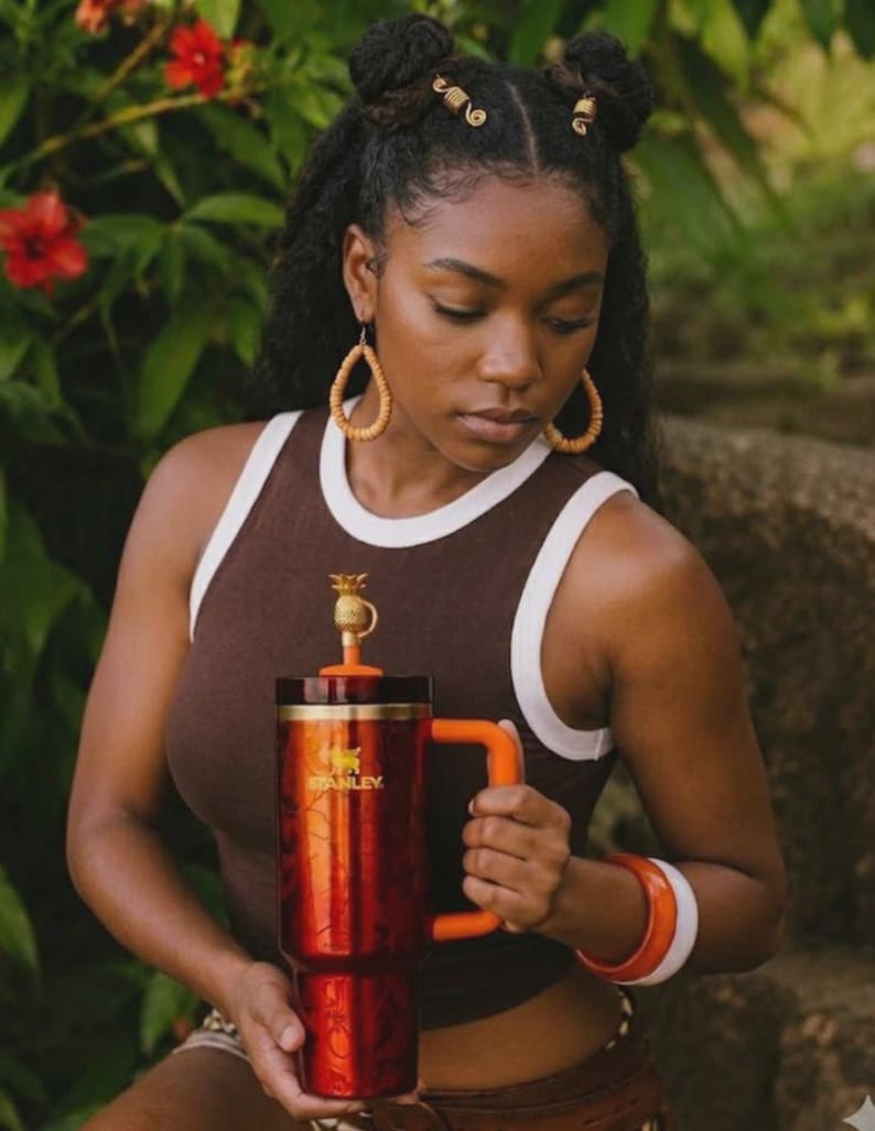 May include: A large, red and orange Stanley travel mug with a pineapple-shaped topper. The mug has a handle and the Stanley logo. The person in the image is wearing a brown tank top with white trim and orange accessories.