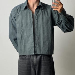 Puede incluir: Una camisa de manga larga abotonada en color verde azulado oscuro, con un sutil patrón de rayas verticales, cuello clásico y un bolsillo en el pecho. La camisa se lleva sobre una camiseta blanca y se combina con vaqueros de mezclilla gris oscuro.