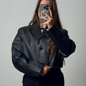 May include: A black cropped blazer with a single front button closure, structured shoulders, and long sleeves. The jacket features a smooth finish, a classic notched lapel collar, and a straight hemline that sits above the waist, worn over a striped shirt.