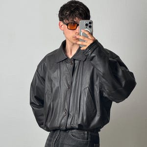 May include: A black leather jacket featuring a classic collar, a button-down front closure, and two side pockets. The jacket has a relaxed, slightly oversized fit with long sleeves and a hem that sits at the waist, paired with dark denim jeans.